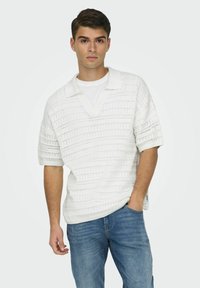 Hvit strikket polo skjorte med korte ermer, med v-hals og åpen vevemønster. Båret over en enkel hvit t-skjorte og parret med blå jeans.