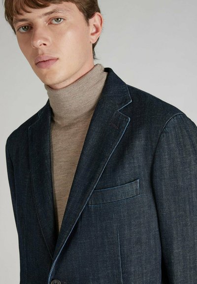 Granatowa dżinsowa marynarka z klapami, pojedynczą kieszenią na piersi i widocznym przeszyciem. Nosi się ją na jasnobeżowym swetrze z golfem.