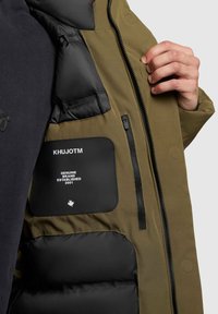 Olivengrøn jakke med en puffet sort inderfor, lynlåslommer og et sort mærke med hvid tekst. Knappedetaljer på ydersiden.