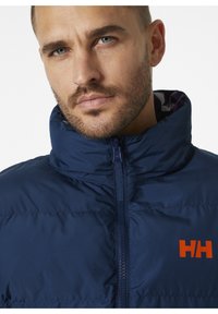 Chaqueta de plumas azul marino con un cuello alto, cremallera frontal y un logo naranja en el lado izquierdo del pecho. Material suave y resistente al agua, diseño acolchado.