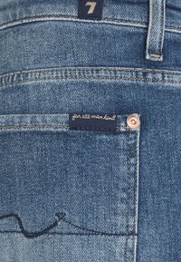 Jeans de mezclilla azules con un bolsillo trasero que tiene una etiqueta que dice "para toda la humanidad", acompañados de costuras visibles y un remache de metal.