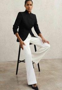 Must värvitud ülikondjakku kõrge kraega ja ühe nööbiga, kombineeritud valgete laia säärega pükstega ja terava nina mustade kontsakingadega.