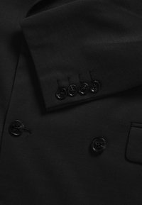 Manche de veste de costume noire avec quatre boutons noirs au-dessus de deux boutons noirs plus grands sur le devant de la veste et une petite poche visible.