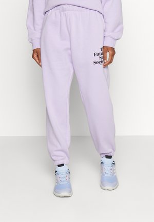 Henkilö pukeutuneena vaaleanpurppuriin collegehousuihin, joissa musta teksti oikeassa reisessä, ja vaaleansinisiin ja valkoisiin lenkkareihin yksivärisellä taustalla.