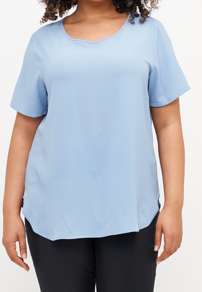 Personne portant un haut texturé bleu clair à manches courtes avec un col arrondi et un pantalon noir, debout devant un fond uni.