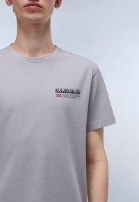 Šviesiai pilkas medvilninis marškinėlis trumpomis rankovėmis, su mažu išsiuvinėtų logotipu kairėje krūtinės pusėje ir matomomis siuvimo detalėmis.