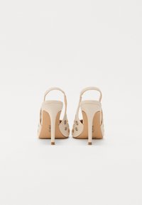Beige high-heeled sandals with an open back, decorative studs, narrow straps, and stiletto heels. Smooth material with a glossy finish.