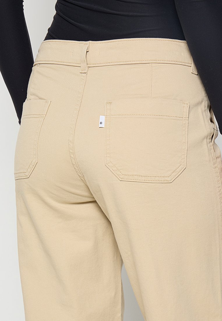 Pantalones de tiro alto beige con dos bolsillos traseros. La tela tiene una textura suave y presenta costuras dobles en los bordes.