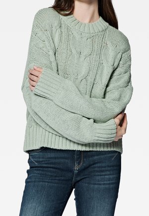 Maglione lavorato a maglia verde chiaro con scollo tondo, motivo a trecce e polsini a coste. Indossato sopra jeans blu scuro.
