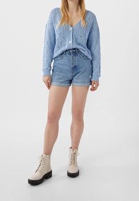 Světlá modrá pletená košile s výstřihem do V a knoflíky, doplněná džínovými šortkami. Krémové šněrovací boty s masivními černými podrážkami.