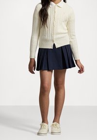 Dívka oblečená v krémovém kardiganu s copánkovým vzorem, námořnické plisované sukni a bílých teniskách, stojící na pozadí s jednoduchým bílým pozadím.