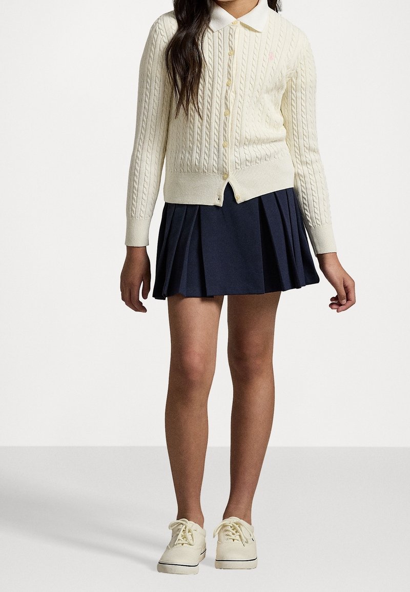 Dívka oblečená v krémovém kardiganu s copánkovým vzorem, námořnické plisované sukni a bílých teniskách, stojící na pozadí s jednoduchým bílým pozadím.