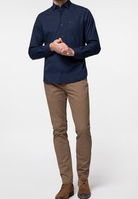 Homme portant une chemise bleu marine à boutons, un pantalon ajusté beige et des chaussures en cuir marron, debout les mains jointes.