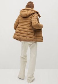 Brun puffer-jakke med vandret syninger og hætte. Jakken har en afslappet pasform og ribkanter, som er parret med lysfarvede bukser.