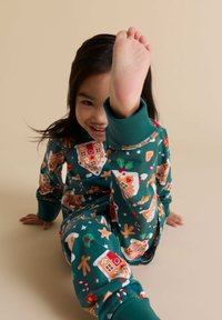 Vihreät puuvillapijamat, joissa on piparkakkutaloja ja juhlakauden kuvioita. Ne ovat rentoa mitoitusta, kuvetta hihansuissa ja leikkisä asento.