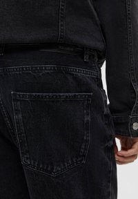 Gros plan sur une personne portant un jean en denim noir et une veste en denim noir, avec un focus sur la poche arrière et les détails latéraux.
