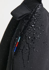 Nærbillede af sort stof med vanddråber og en lille syet detalje med blå, røde og lyseblå lodrette striber.