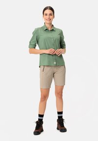 Camicia verde chiaro, con bottoni e maniche arrotolate, abbinata a pantaloni shorts beige con tasche con zip. Stivali da trekking marroni e calze a righe nere.