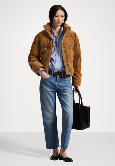 Jachetă puffer din catifea închisă la culoare, cu guler, prevăzută cu buzunare frontale. Cămașă din denim deschis la culoare, blugi albaștri, geantă tote neagră și pantofi fără toc negri.