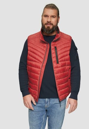 Rote gesteppte Puffer-Weste mit hohem Kragen, Reißverschluss und einer schwarzen Akzenttasche; getragen über einem marineblauen strukturierten Pullover, kombiniert mit hellblauen Jeans.