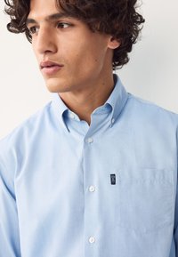 Camicia azzurro chiaro in cotone con bottoni, caratterizzata da una tasca frontale e una piccola etichetta con il marchio sul petto sinistro. Tessuto liscio, colletto standard.