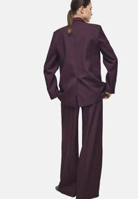 Costume bordeaux sur mesure avec un blazer structuré et un pantalon à jambes larges. Tissu lisse avec une silhouette classique et une fente au dos.