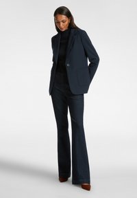 Granatowa marynarka z jednym guzikiem, dwiema przednimi kieszeniami i dopasowanym krojem, zestawiona z ciemnymi dzwonami i brązowymi szpilkami.