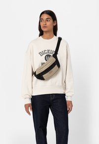 Borsa a tracolla beige e nera con due scomparti con cerniera, materiale in tessuto e tracolla regolabile, indossata da una persona in felpa bianca.