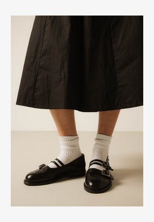 Picioare inferioare purtând șosete albe și pantofi Mary Jane negri cu cataramă și perforații decorative, sub o fustă neagră de lungime medie, pe un fundal simplu.