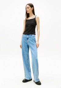 Top de tirantes negro acanalado con borde de encaje, combinado con jeans de pierna ancha en azul claro. La modelo lleva zapatos de plataforma negros. Fondo sencillo.