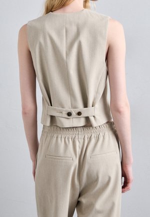 Persona que lleva un chaleco beige sin mangas con detalle de correa con botones en la espalda y pantalones beige a juego con cintura elástica.