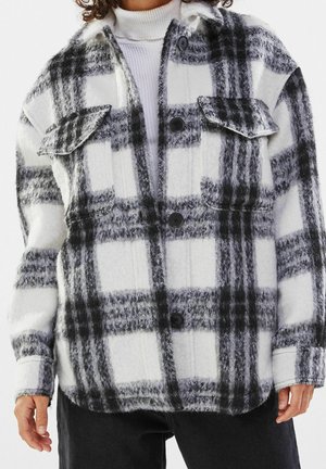 Person trägt eine schwarz-weiß karierte, flauschige Hemdjacke über einem weißen Rollkragenpullover und schwarzen Hosen.