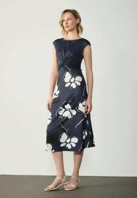 Frau steht und trägt ein marineblaues Midikleid mit großen weißen Blumenmustern und beigen Sandalen mit Riemchen.