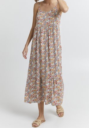 Frau trägt ein ärmelloses Midi-Kleid mit floralem Muster, dünnen Trägern und beigen flachen Sandalen vor einfarbigem Hintergrund.