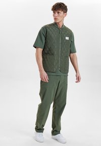 Prošívaná zelená vesta s vysokým límcem, zipem a dvěma předními kapsami, nošená přes zelenou košili a spárovaná s odpovídajícími kalhotami.