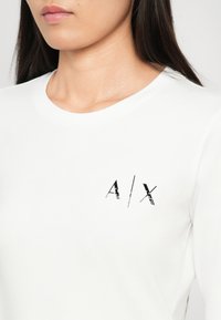 Hvit crew neck t-skjorte med en tettsittende design som har en svart grafisk "A/I/X" logo på venstre brystområde. Glatt bomullsstoff.