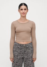 Jeune femme aux cheveux bruns portant un crop top beige à manches longues et un pantalon gris à imprimé zèbre, se tenant devant un fond blanc uni.
