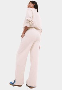 Beige sweatshirt en bijpassende wijde broek, beide gemaakt van zachte stof. De broek heeft zijzakken en sleuven aan de onderkant, gedragen met blauwe schoenen.