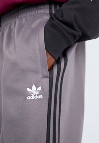 Grå treningbukser med en glatt tekstur, med svarte sidestriper og en hvit Adidas-logo. Elastisk liv med sidelommer.