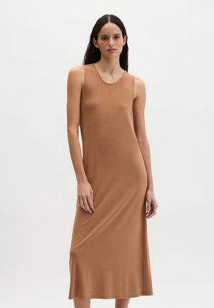 Kobieta o długich ciemnych włosach ubrana w bezrękawową, beżową suknię sięgającą kostek, stojąca na tle jasnego, gładkiego tła.
