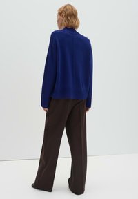 Pull en maille bleue avec col montant, poignets et ourlet côtelés. Associé à un pantalon marron à jambes larges, présenté sur un fond clair.