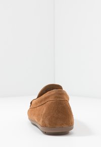 Chaussure de style mocassin en suède marron avec un profil bas, un talon arrondi et une surface texturée. Présente un contour cousu le long de la couture.