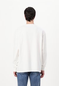 Long-sleeve white cotton t-shirt with a crew neckline, relaxed fit, and straight hem. Worn with light blue denim jeans.
