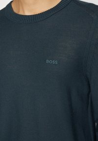 Gros plan sur un pull en maille bleu sarcelle foncé avec un col rond côtelé et "BOSS" brodé sur la poitrine.
