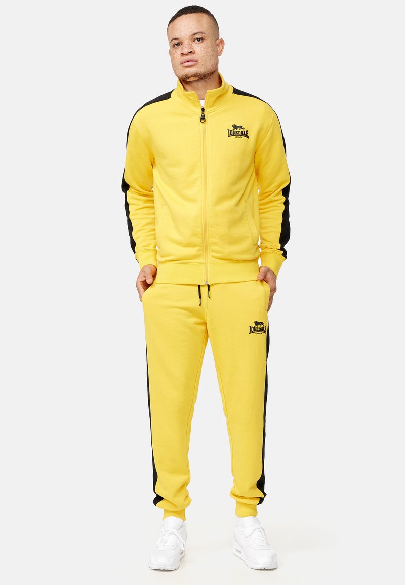 Žltý zip-up športový overal s čiernymi bočnými panelmi a logom. Má stojatý golier a manžety; vyrobený z jemného materiálu. Biely tenisky dopĺňajú vzhľad.
