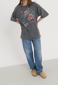 Szary, oversized t-shirt z kwiatowym nadrukiem w kolorach różowym i tealowym. W zestawie z jasnoniebieskimi dżinsami i beżowymi klapkami. Prosty, casualowy design.