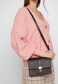 Borsa a tracolla in pelle nera testurizzata con patta e chiusura metallica dorata, tenuta accanto a un modello che indossa un maglione di maglia rosa e pantaloni a quadri.