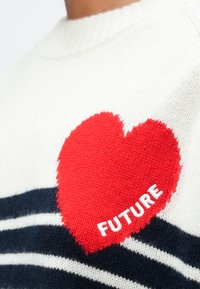 Suéter blanco de punto con rayas negras y un parche en forma de corazón rojo bordado con la palabra "FUTURE" en el pecho.