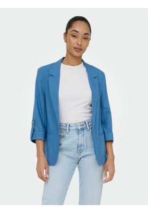 Žena v modrom sakom s vyhrnutými rukávmi cez biele tričko a svetlomodré rifle, stojaca pred jednoduchým bielym pozadím.