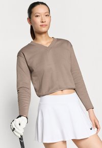 Καφέ, κοντό μακρυμάνικο τοπ με V-λαιμόκοψη, συνδυασμένο με μια λευκή φούστα με δακτύλιο. Λευκό γάντι κρατάει μια ρακέτα γκολφ.